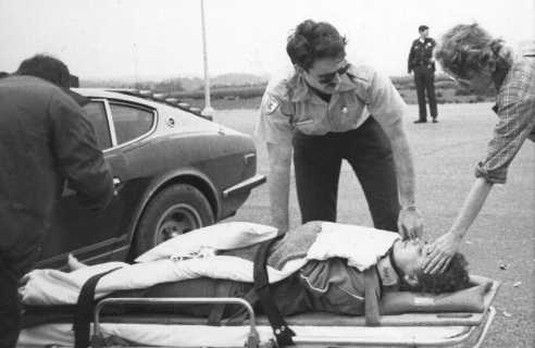A child lies strapped to a stretcher with a neck brace as two adults attend to them. A uniformed officer stands nearby, and an older car is parked in the background.