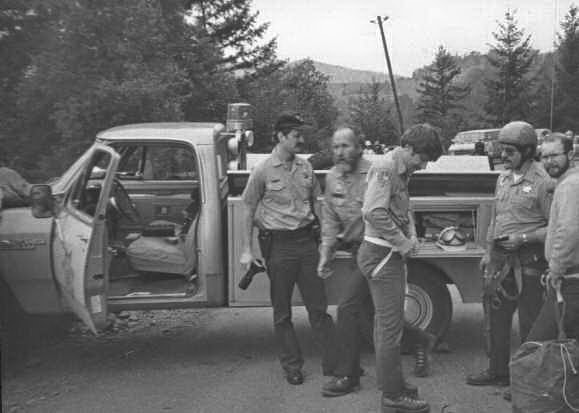 Four uniformed men stand beside an open pickup truck in a wooded area, talking and handling equipment. The setting appears rural with trees and hills in the background.