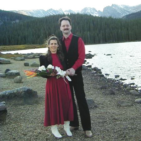A smiling couple stands by a lakeshore with mountains and trees in the background. The woman wears a red dress and white boots, holding a bouquet, while the man wears a red shirt, black vest, and pants.