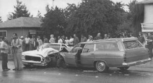 Two damaged cars, a sedan and a station wagon, have collided head-on in the street. A crowd of people stands nearby, observing the scene. The car doors are open and the weather appears overcast.