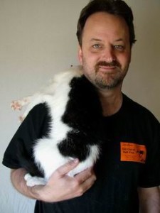 A man with short dark hair and a beard smiles at the camera, while a black and white cat rests on his shoulder with its body and paws draped over him. The man is wearing a black shirt with an orange name tag.