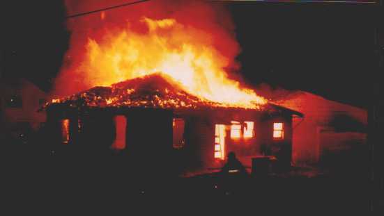 A house engulfed in flames at night, with a large fire consuming the roof and bright orange light illuminating the scene. Silhouettes and neighboring structures are visible in the background.