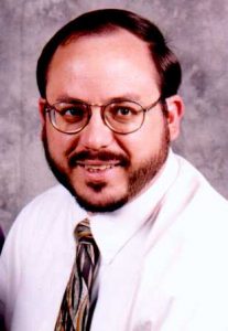 A man with short brown hair, glasses, and a beard, wearing a white shirt and patterned tie, smiles at the camera against a gray mottled background.