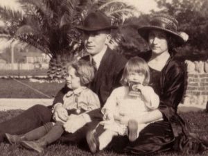 A sepia-toned photo of a man and woman sitting on grass with two young children, one sitting on each of their laps. Palm trees and a stone wall are visible in the background.