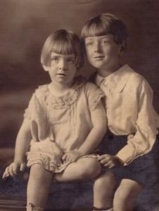 Two young children in vintage clothing sit closely together, posing for a sepia-toned portrait. The younger child wears a light dress, and the older child wears a button-up shirt and shorts. Both have short hair and neutral expressions.