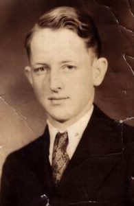 A sepia-toned portrait of a young man with neatly combed hair, wearing a suit, dress shirt, and tie, looking slightly to the side with a neutral expression. The photo has visible creases and wear.