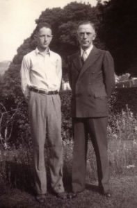Two men stand side by side in a garden. The younger man on the left wears glasses, a light shirt, and trousers, while the older man on the right wears a double-breasted suit. Trees and bushes are in the background.