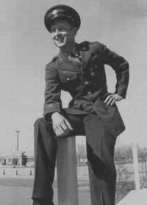 A young man in a military uniform and cap poses outdoors, smiling and resting one hand on his knee. The background features trees and a building under a clear sky.
