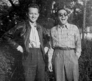 Two men stand outdoors, smiling and posing for the camera. One wears a leather jacket with his hand on his hip, while the other, in glasses and a plaid shirt, stands laughing. Trees and foliage are visible in the background.