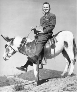 A smiling man in a suit sits sideways on a mule, holding a book and resting one foot on the ground. The photo is black and white with hills visible in the background.