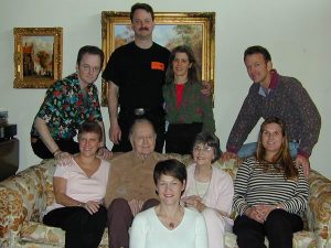 Nine adults pose for a group photo indoors, with three men and one woman standing behind a couch, and five people seated, including two elderly individuals. Framed paintings decorate the wall behind them.