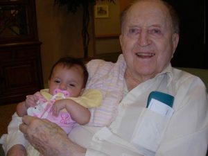 An elderly man smiles while holding a baby with a pink pacifier. The baby is dressed in light clothing and lies comfortably in the mans arms. Papers are visible in the mans shirt pocket.