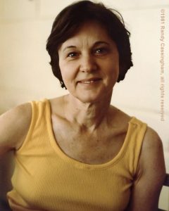 A woman with short dark hair, wearing a yellow sleeveless top, smiles gently at the camera. The background is softly lit and neutral. ©1981 Randy Cassingham, all rights reserved.