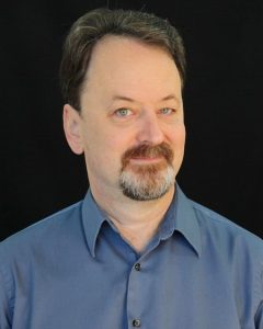 A middle-aged man with light skin, short brown hair, and a graying goatee is wearing a blue collared shirt and posing in front of a plain black background. He is looking slightly to the side and smiling softly.
