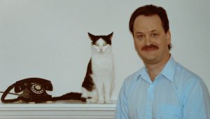 A man with a mustache in a light blue shirt stands beside a black-and-white cat sitting on a shelf near a black rotary telephone, against a plain white background.