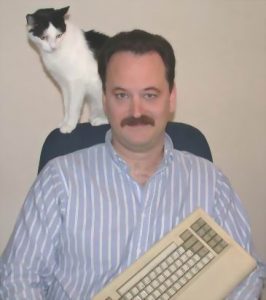 A man with a mustache sits in a chair holding a computer keyboard, while a black-and-white cat stands on the back of the chair, looking down at him.