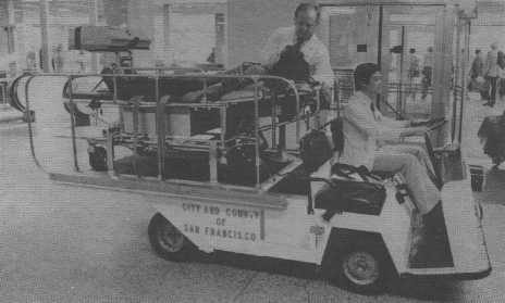A black and white photo shows a medical transport cart marked City and County of San Francisco carrying a patient on a stretcher with two attendants in an indoor public space, possibly an airport or hospital.