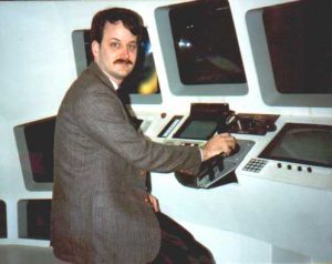A man with dark hair and a mustache, wearing a suit, sits at a control panel with multiple screens and buttons, looking at the camera.