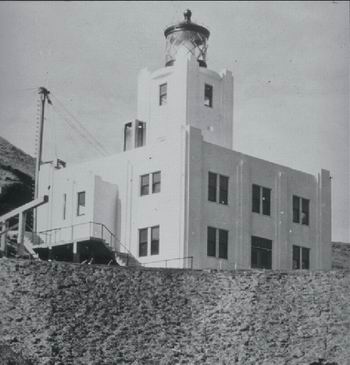 Black and white photo of a lighthouse with a square, white stucco building and a lantern room on top, situated on a rocky hill with visible stairs and railings leading to the entrance.