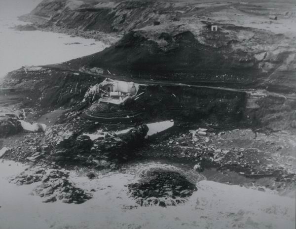 Black and white aerial view of a coastal landslide with debris scattered at the base of a cliff; a damaged structure is partially buried, and rough terrain leads to the shoreline.